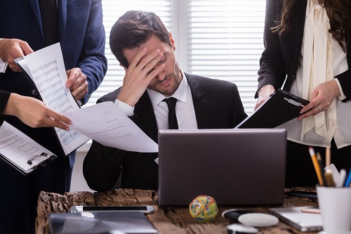 Stress and Employment Law Image shows caucasian man in a black suit and tie, under stress, at a desk with a laptop surrounded by people with papers, seemingly to hassle him