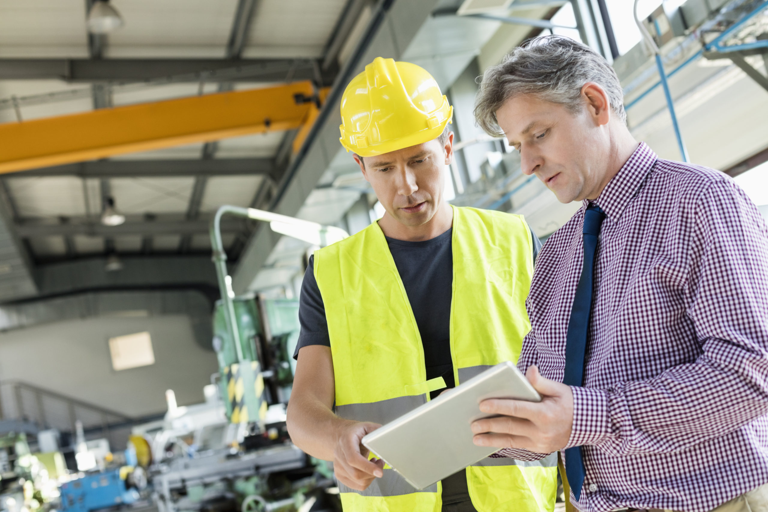 The Benefits of Outsourcing Health and Safety displayed by a worker and manager in a warehouse discussing plans over a tablet. The worker is wearing high visibility PPE and a hard hat.